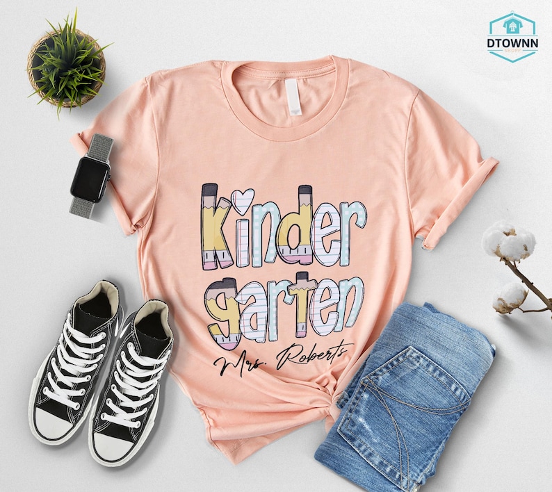 K&ouml;nnte beinhalten: Pfirsichfarbenes T-Shirt mit dem Wort "Kindergarten" in einem farbenfrohen Design im Bleistift-Stil. Der Name "Mrs. Roberts" ist darunter aufgedruckt. Das Shirt wird mit blauen Jeans und schwarzen Turnschuhen kombiniert.