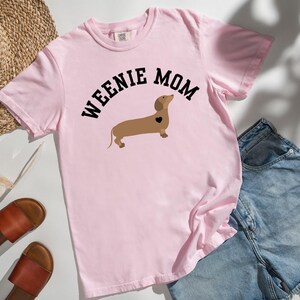 May include: A light pink t-shirt with the words "WEENIE MOM" arched above a brown dachshund illustration with a black heart. The shirt is paired with denim shorts, sandals, and a straw hat.