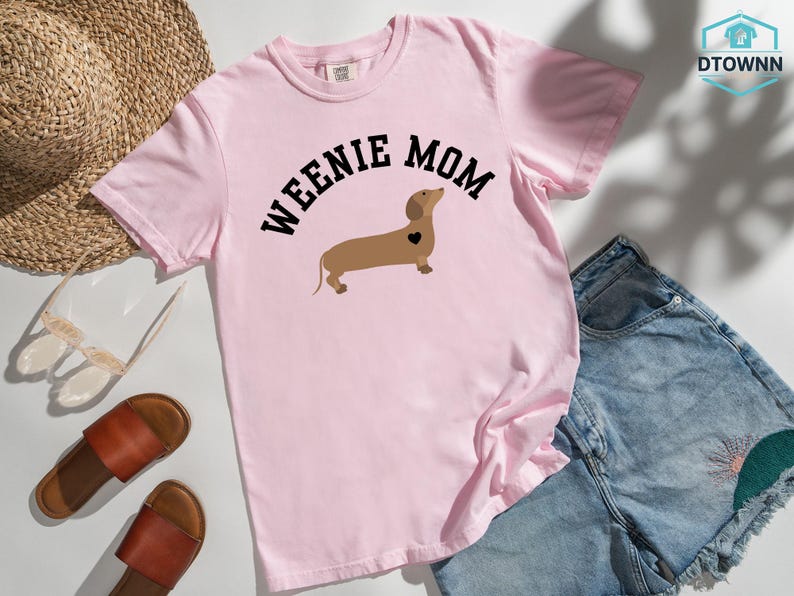 May include: A light pink t-shirt with the words "WEENIE MOM" arched above a brown dachshund illustration with a black heart. The shirt is paired with denim shorts, sandals, and a straw hat.
