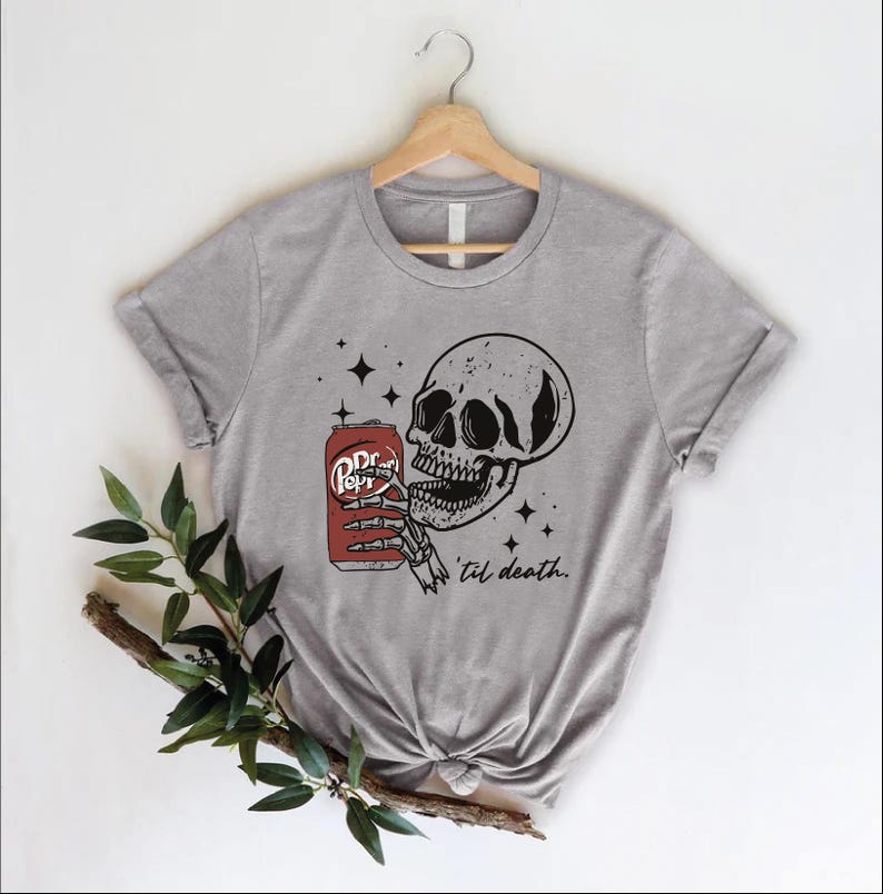 Op de afbeelding: Een grijs gem&ecirc;leerd T-shirt met korte mouwen en een zwart-witte afbeelding van een menselijke schedel die een rood frisdrankblikje met het label Pepper vasthoudt. Het ontwerp bevat kleine zwarte sterren en de cursieve tekst 'til death. Het shirt hangt aan een hanger.