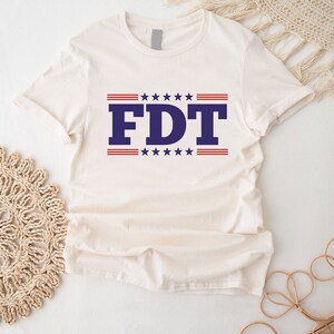 May include: A white t-shirt with a blue and red design. The design features the letters "FDT" in blue with red and white stars above and below the letters.