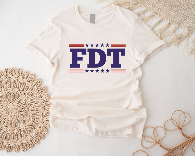 May include: A white t-shirt with a blue and red design. The design features the letters "FDT" in blue with red and white stars above and below the letters.