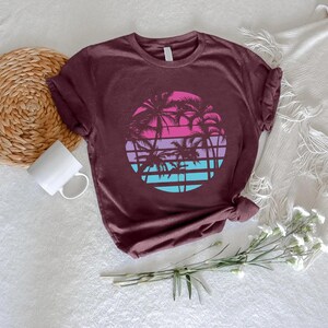 May include: A maroon t-shirt featuring a circular graphic of palm trees against a sunset. The sunset design includes bands of pink, purple, and blue. The shirt is made of a soft material and has a crew neck.