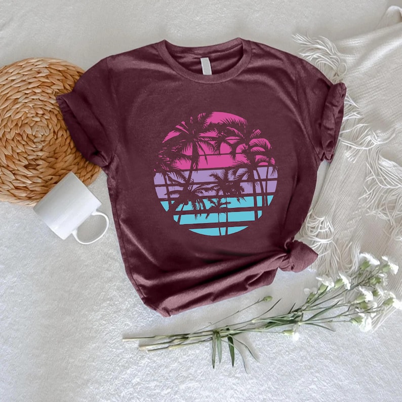 May include: A maroon t-shirt featuring a circular graphic of palm trees against a sunset. The sunset design includes bands of pink, purple, and blue. The shirt is made of a soft material and has a crew neck.