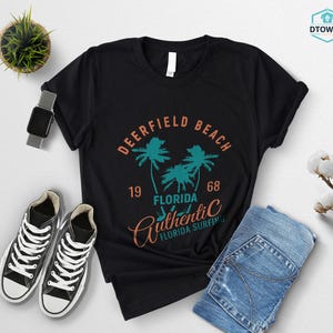 May include: Black t-shirt with a Deerfield Beach, Florida graphic. The design includes palm trees, the year 1968, and the words "Authentic Florida Surfing". The shirt is paired with blue jeans and black sneakers.