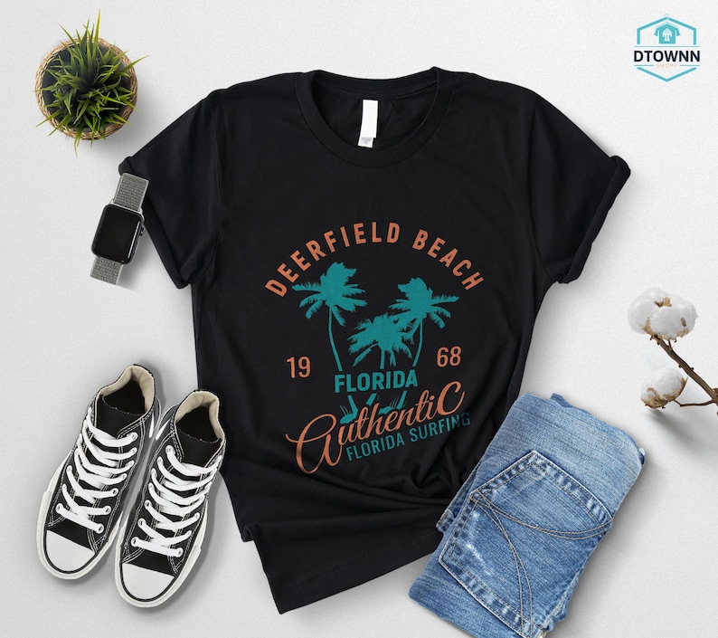 May include: Black t-shirt with a Deerfield Beach, Florida graphic. The design includes palm trees, the year 1968, and the words "Authentic Florida Surfing". The shirt is paired with blue jeans and black sneakers.