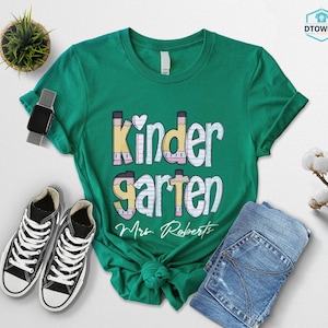 K&ouml;nnte beinhalten: Ein gr&uuml;nes T-Shirt mit dem Wort "Kindergarten" in einer verspielten Schriftart, mit Bleistift- und Herzdetails. Das Shirt enth&auml;lt auch den Namen "Mrs. Roberts" in Schreibschrift. Schwarz-wei&szlig;e Turnschuhe und eine blaue Jeans liegen neben dem Shirt.