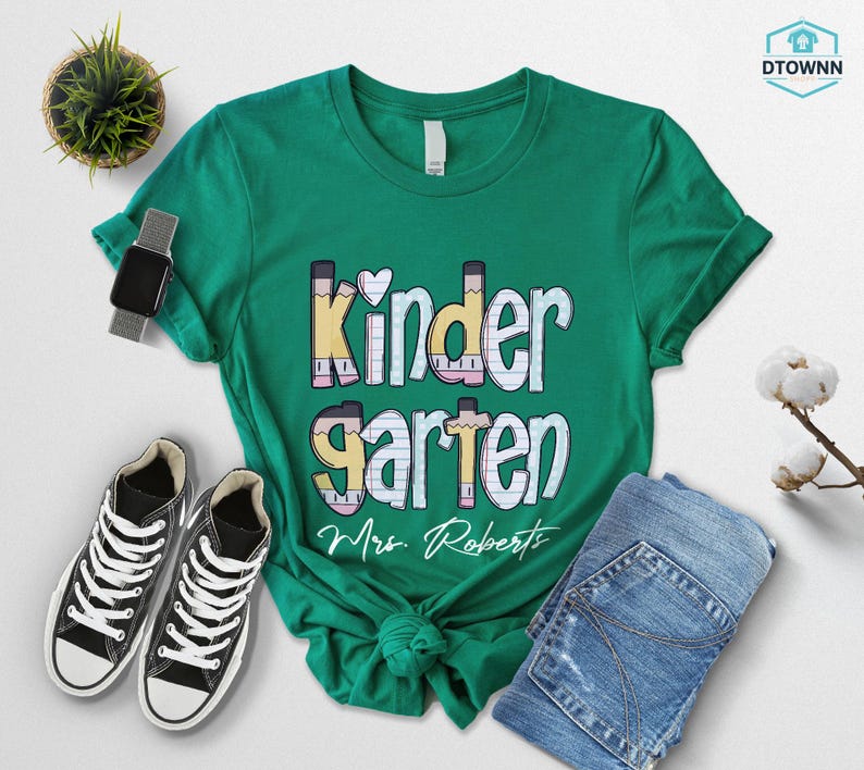 K&ouml;nnte beinhalten: Ein gr&uuml;nes T-Shirt mit dem Wort "Kindergarten" in einer verspielten Schriftart, mit Bleistift- und Herzdetails. Das Shirt enth&auml;lt auch den Namen "Mrs. Roberts" in Schreibschrift. Schwarz-wei&szlig;e Turnschuhe und eine blaue Jeans liegen neben dem Shirt.