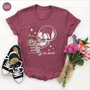 Op de afbeelding: Een heather maroon t-shirt met korte mouwen en een witte afbeelding van een skeletthand die een frisdrankblikje vasthoudt met de tekst Pepper en de zin 'til death. Het ontwerp bevat kleine steraccenten rondom de schedel en de hand.