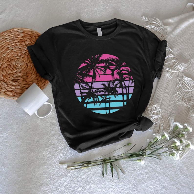 May include: Black t-shirt featuring a circular graphic of palm trees against a gradient sunset in pink, purple, and blue. The design includes horizontal lines and silhouettes of palm trees, evoking a tropical theme.