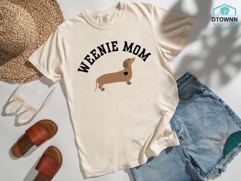 May include: Cream-colored t-shirt with the text "WEENIE MOM" above a brown dachshund illustration with a black heart. The shirt is paired with denim shorts, sandals, and a straw hat.