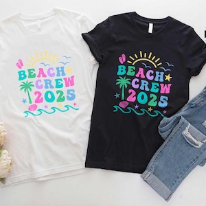 Puede incluir: Dos camisetas, una blanca y otra negra, con un dise&ntilde;o colorido de "Beach Crew 2025". El dise&ntilde;o incluye un sol, una palmera, olas y estrellas. Un par de vaqueros azules est&aacute; al lado de la camiseta negra.