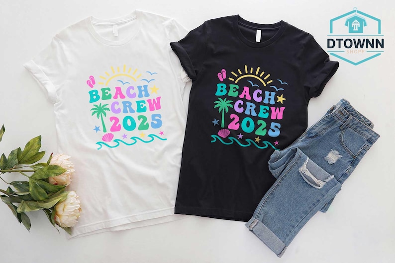 Puede incluir: Dos camisetas, una blanca y otra negra, con un dise&ntilde;o colorido de "Beach Crew 2025". El dise&ntilde;o incluye un sol, una palmera, olas y estrellas. Un par de vaqueros azules est&aacute; al lado de la camiseta negra.