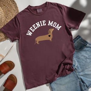 May include: Burgundy t-shirt with the text "WEENIE MOM" in white, above a brown dachshund illustration with a black heart. The shirt is paired with denim shorts, sandals, and a straw hat.