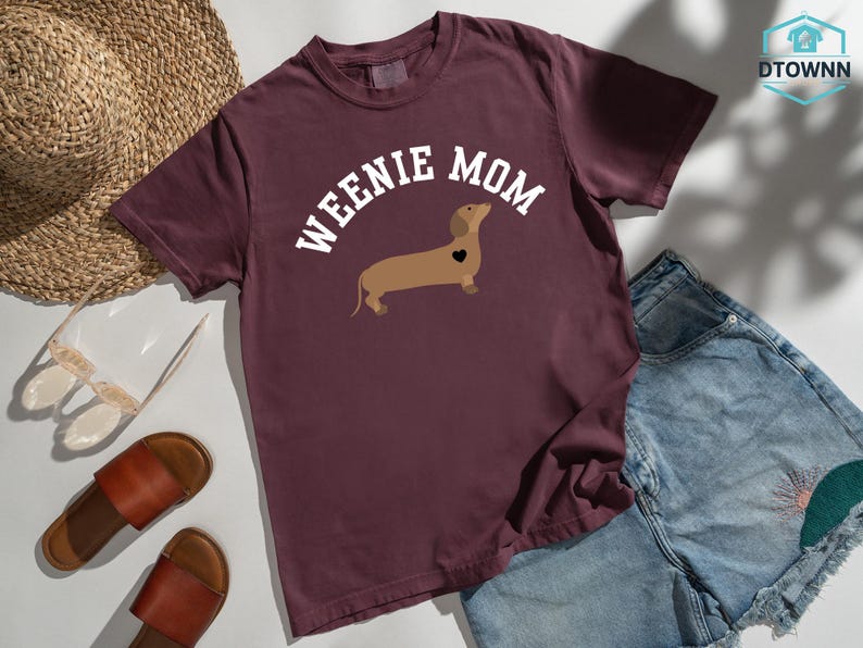 May include: Burgundy t-shirt with the text "WEENIE MOM" in white, above a brown dachshund illustration with a black heart. The shirt is paired with denim shorts, sandals, and a straw hat.