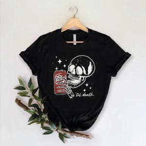 Op de afbeelding: Een zwart t-shirt met korte mouwen en een witte afbeelding van een skeletthand die een rood frisdrankblikje vasthoudt. Het ontwerp bevat vierpuntige sterren en de cursieve tekst 'til death.' Het t-shirt hangt aan een houten kledinghanger.