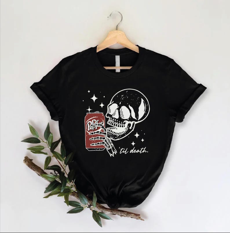 Op de afbeelding: Een zwart t-shirt met korte mouwen en een witte afbeelding van een skeletthand die een rood frisdrankblikje vasthoudt. Het ontwerp bevat vierpuntige sterren en de cursieve tekst 'til death.' Het t-shirt hangt aan een houten kledinghanger.