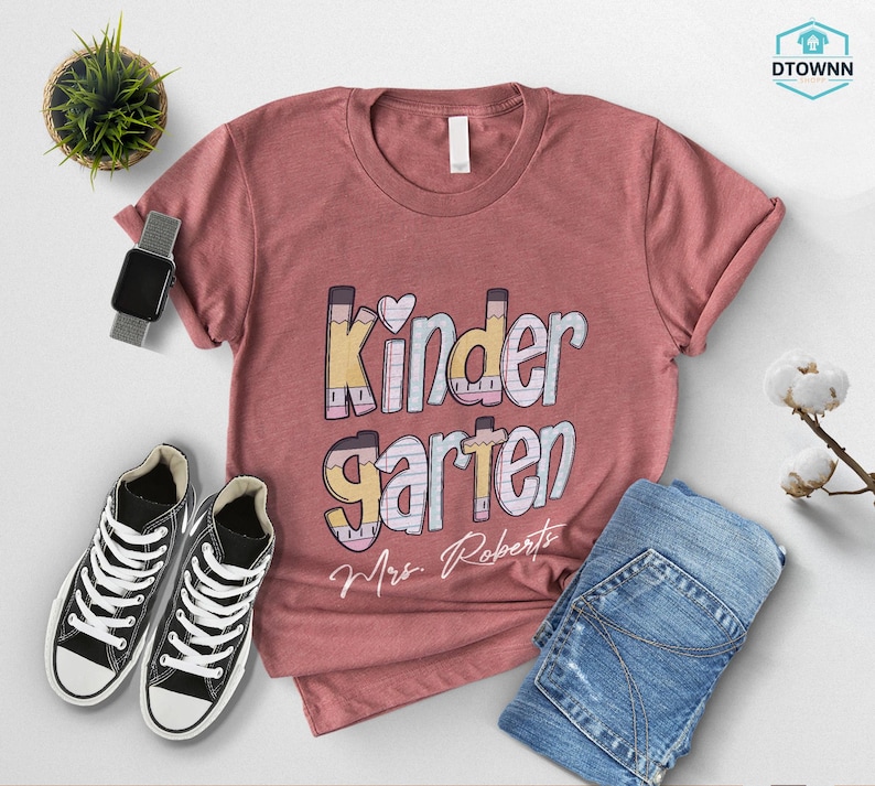 K&ouml;nnte beinhalten: Ein staubrosa T-Shirt mit dem Wort "Kindergarten" in einer farbenfrohen, verspielten Schriftart. Das Shirt zeigt auch ein Herz- und Bleistift-Design sowie den Namen "Mrs. Roberts". Das Shirt wird mit einer blauen Jeans und schwarzen Turnschuhen kombiniert.