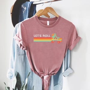 Puede incluir: Camiseta rosa polvoriento con el texto "LET'S ROLL" encima de un gr&aacute;fico de patines retro. El gr&aacute;fico presenta una franja de arco&iacute;ris colorida y un dise&ntilde;o de patines en turquesa, naranja y rojo. La camiseta est&aacute; colgada en una percha de madera.