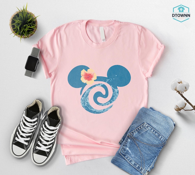 May include: A light pink t-shirt featuring a blue Mickey Mouse silhouette with a floral design. The shirt is paired with black sneakers, blue jeans, and a smartwatch. A small potted plant and cotton sprig are also visible.