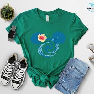 May include: A green t-shirt featuring a blue Mickey Mouse silhouette with a yellow and red flower and a water design. The shirt is paired with blue jeans and black sneakers with white laces.