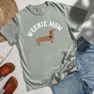May include: A light gray t-shirt with the words "WEENIE MOM" above a brown dachshund with a black heart. The shirt is paired with denim shorts, brown sandals, and a straw hat.