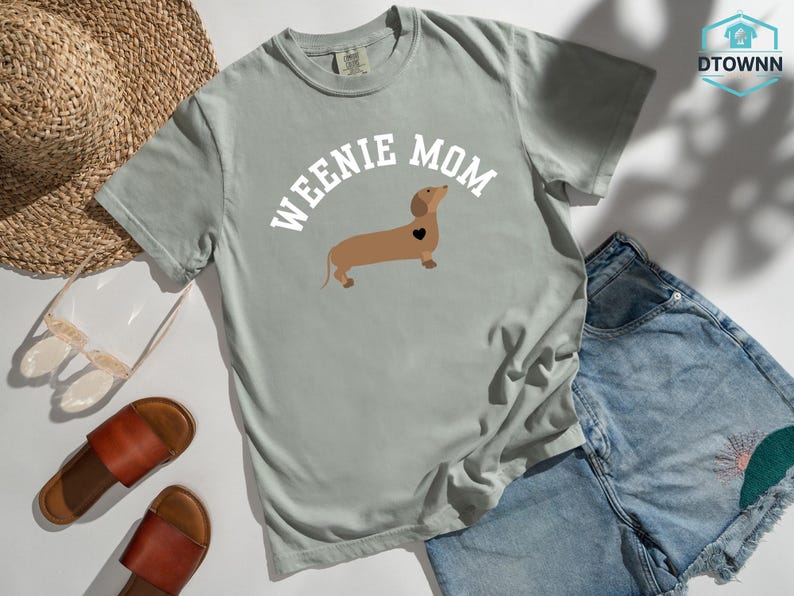 May include: A light gray t-shirt with the words "WEENIE MOM" above a brown dachshund with a black heart. The shirt is paired with denim shorts, brown sandals, and a straw hat.