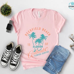 May include: A light pink t-shirt with a graphic that reads "Deerfield Beach Florida Authentic Surfing 1968" in teal and orange. The shirt is paired with black sneakers, blue jeans, and a smartwatch.