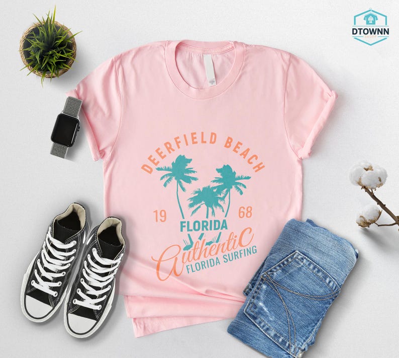 May include: A light pink t-shirt with a graphic that reads "Deerfield Beach Florida Authentic Surfing 1968" in teal and orange. The shirt is paired with black sneakers, blue jeans, and a smartwatch.