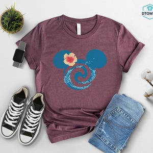 May include: A maroon t-shirt featuring a blue Mickey Mouse head design with a floral accent and a wave pattern. The shirt is paired with blue jeans, black sneakers, and a smartwatch. A small potted plant and cotton stems are also visible.
