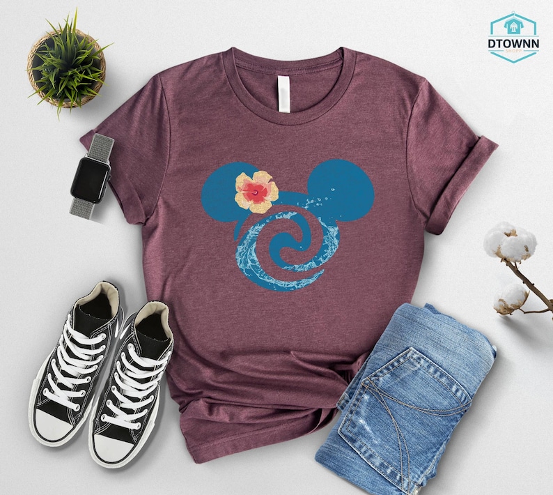 May include: A maroon t-shirt featuring a blue Mickey Mouse head design with a floral accent and a wave pattern. The shirt is paired with blue jeans, black sneakers, and a smartwatch. A small potted plant and cotton stems are also visible.