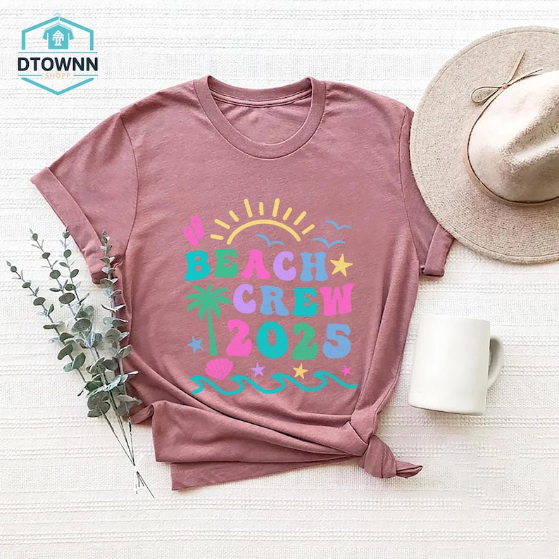 Puede incluir: Camiseta malva con la frase "BEACH CREW 2025" en letras coloridas y divertidas. El dise&ntilde;o incluye un sol, una palmera, olas y estrellas. Se ven un sombrero beige y una taza blanca.