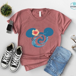 May include: A dusty rose t-shirt featuring a blue Mickey Mouse silhouette with a floral accent and a wave design. The shirt is paired with blue jeans and black sneakers. A smartwatch and a small potted plant are also visible.