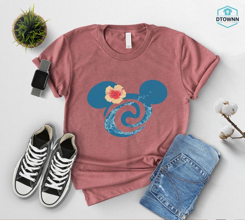 May include: A dusty rose t-shirt featuring a blue Mickey Mouse silhouette with a floral accent and a wave design. The shirt is paired with blue jeans and black sneakers. A smartwatch and a small potted plant are also visible.