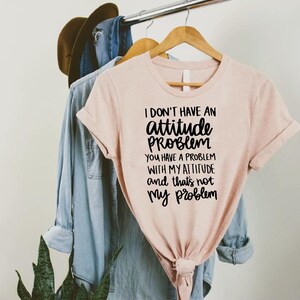 K&ouml;nnte beinhalten: Hellrosa T-Shirt mit schwarzem Text: "I don't have an attitude problem, you have a problem with my attitude and that's not my problem". Das Shirt h&auml;ngt an einem Holzb&uuml;gel mit anderer Kleidung.