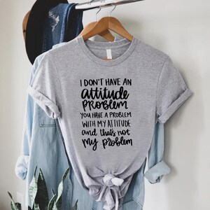K&ouml;nnte beinhalten: Ein meliertes graues T-Shirt mit dem Text "I DON'T HAVE AN attitude PROBLEM YOU HAVE A PROBLEM WITH MY ATTITUDE and that's not my problem" in schwarzer Schrift. Das T-Shirt h&auml;ngt an einem Holzb&uuml;gel.