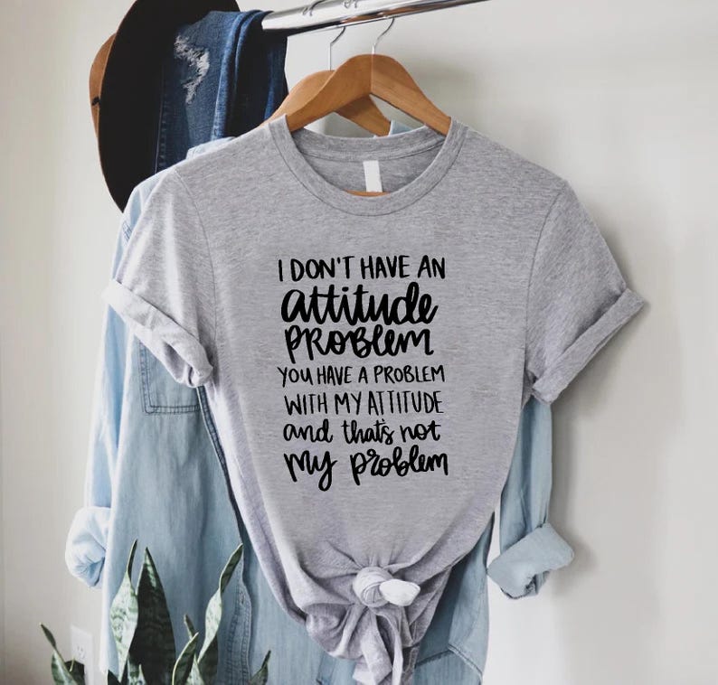 K&ouml;nnte beinhalten: Ein meliertes graues T-Shirt mit dem Text "I DON'T HAVE AN attitude PROBLEM YOU HAVE A PROBLEM WITH MY ATTITUDE and that's not my problem" in schwarzer Schrift. Das T-Shirt h&auml;ngt an einem Holzb&uuml;gel.