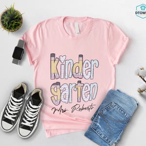 K&ouml;nnte beinhalten: Ein hellrosa T-Shirt mit dem Wort "Kindergarten" in einem farbenfrohen Design mit Bleistiften. Der Name "Mrs. Roberts" steht darunter. Schwarz-wei&szlig;e Turnschuhe und blaue Jeans sind ebenfalls im Bild.