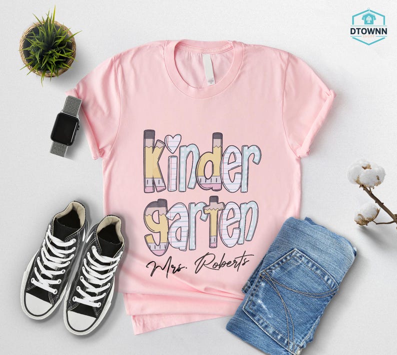 K&ouml;nnte beinhalten: Ein hellrosa T-Shirt mit dem Wort "Kindergarten" in einem farbenfrohen Design mit Bleistiften. Der Name "Mrs. Roberts" steht darunter. Schwarz-wei&szlig;e Turnschuhe und blaue Jeans sind ebenfalls im Bild.