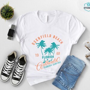 May include: White t-shirt with a graphic design featuring the words "Deerfield Beach Florida" and palm trees in teal and orange. The shirt is paired with black sneakers, blue jeans, and a smartwatch.