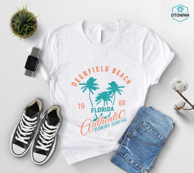 May include: White t-shirt with a graphic design featuring the words "Deerfield Beach Florida" and palm trees in teal and orange. The shirt is paired with black sneakers, blue jeans, and a smartwatch.