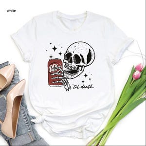 Op de afbeelding: Een wit t-shirt met een zwart-witte afbeelding van een skeletthand die een rood blikje frisdrank vasthoudt. Het ontwerp bevat vierpuntige sterren en de cursieve tekst 'til death.' in zwarte inkt onder de kaaklijn van het skelet.