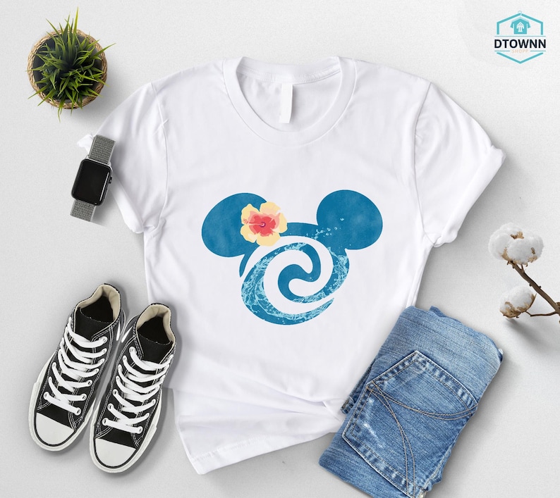 May include: White t-shirt featuring a blue Mickey Mouse head design with a flower and wave pattern. The shirt is paired with black sneakers, blue jeans, and a smartwatch. A small plant and cotton branch are also visible.