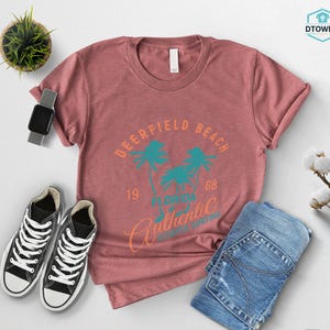 May include: A dusty rose t-shirt with a graphic that reads "Deerfield Beach Florida Authentic Surfing" in teal and orange. The shirt is paired with blue jeans and black and white sneakers.