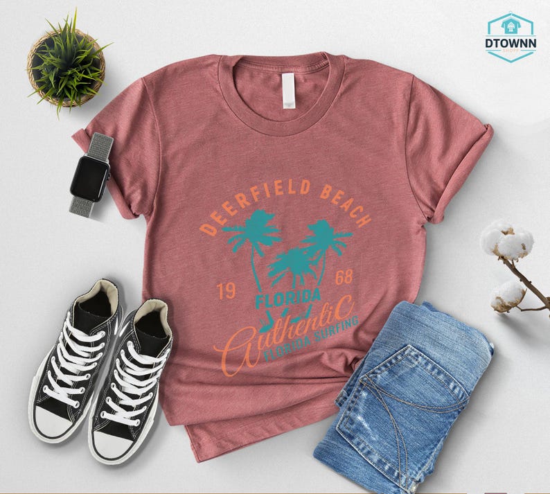 May include: A dusty rose t-shirt with a graphic that reads "Deerfield Beach Florida Authentic Surfing" in teal and orange. The shirt is paired with blue jeans and black and white sneakers.