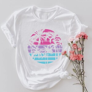 May include: White t-shirt featuring a circular design with palm trees in pink, purple, and blue. The design is set against a grid pattern, evoking a tropical sunset. The shirt is styled with rolled-up sleeves.