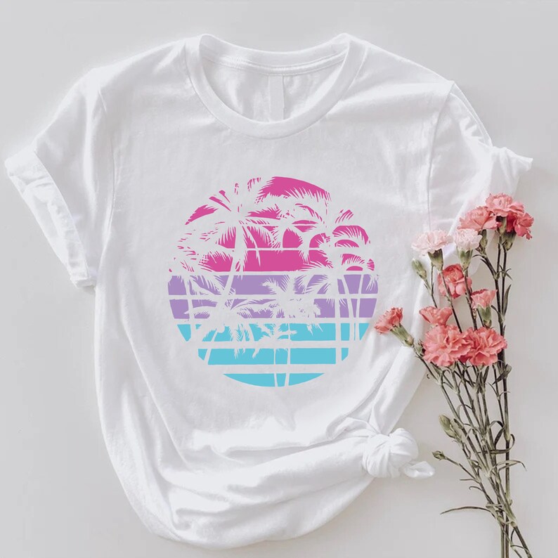 May include: White t-shirt featuring a circular design with palm trees in pink, purple, and blue. The design is set against a grid pattern, evoking a tropical sunset. The shirt is styled with rolled-up sleeves.