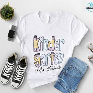 K&ouml;nnte beinhalten: Wei&szlig;es T-Shirt mit dem Wort "Kindergarten" in bunten Buchstaben im Bleistift-Design, einem Herz und dem Namen "Mrs. Roberts" in Schriftform. Schwarze Turnschuhe, eine Smartwatch und eine gefaltete blaue Jeans sind ebenfalls zu sehen.