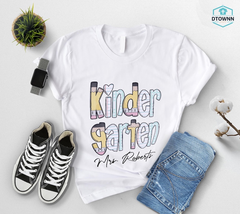 K&ouml;nnte beinhalten: Wei&szlig;es T-Shirt mit dem Wort "Kindergarten" in bunten Buchstaben im Bleistift-Design, einem Herz und dem Namen "Mrs. Roberts" in Schriftform. Schwarze Turnschuhe, eine Smartwatch und eine gefaltete blaue Jeans sind ebenfalls zu sehen.