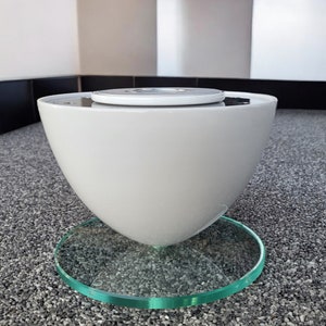 Könnte beinhalten: Eine weiße Keramikschale mit einem runden, silbernen Deckel steht auf einem klaren Glasfuß. Die Schale ist mit Wasser gefüllt und der Fuß steht auf einer grauen, kiesbedeckten Oberfläche.